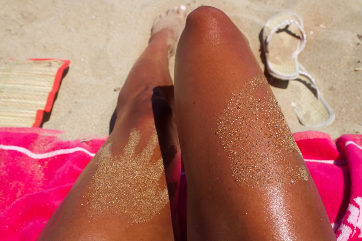 Sun-kissed tanned legs on a pink beach towel, dusted with golden sand